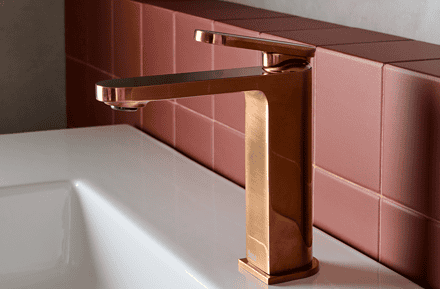 Grifo monomando Roca Kay en acabado cobre cepillado sobre lavabo blanco en baño de diseño contemporáneo con pared de microcemento y azulejos rosas